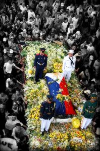 Image of a coffin surrounded by yellow and white flowers with four soldiers standing guard. The crowd surrounding the funeral procession is in black and white.