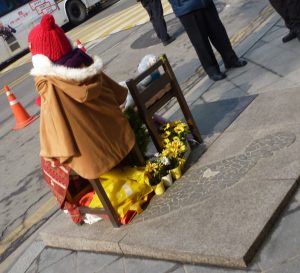 Statue dressed in a brown coat and jacket surrounded by yellow flowers.